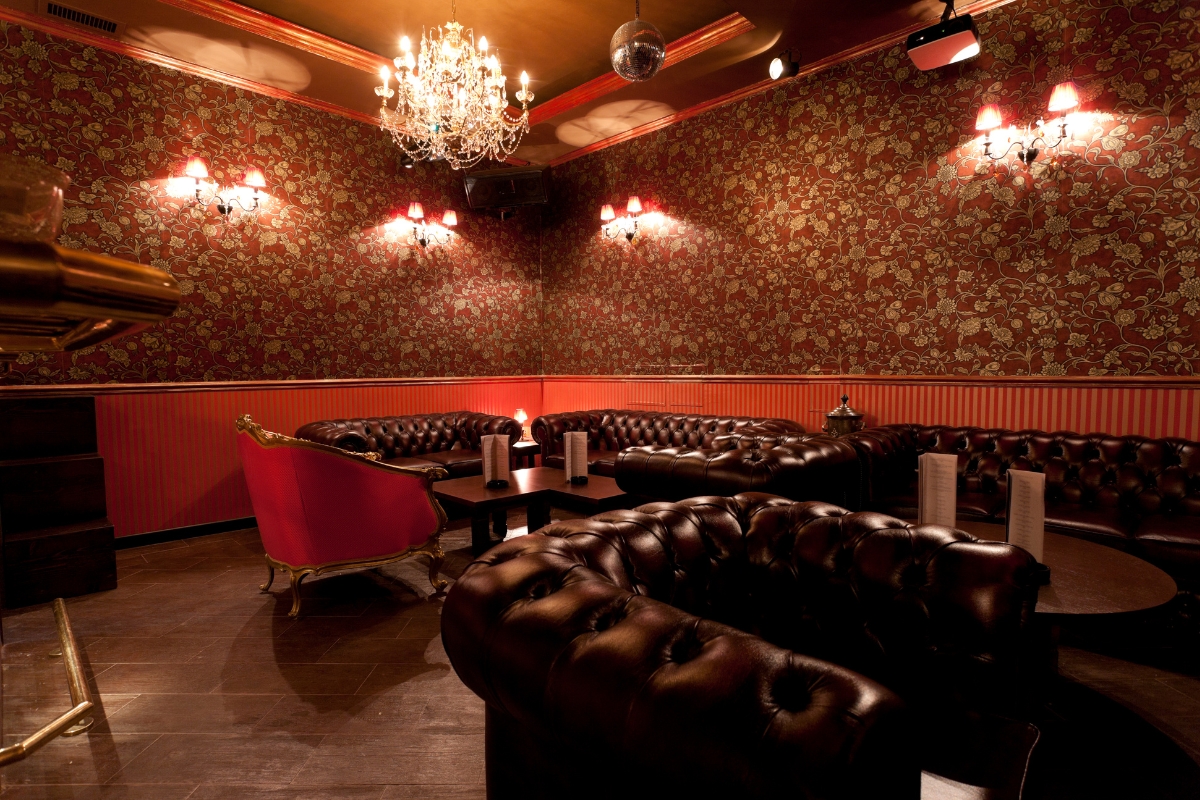 Luxuriously decorated room with dark leather sofas, red patterned wallpaper, ornate chandelier, and ambient wall sconces providing soft light, complemented by subtle cigar decor.