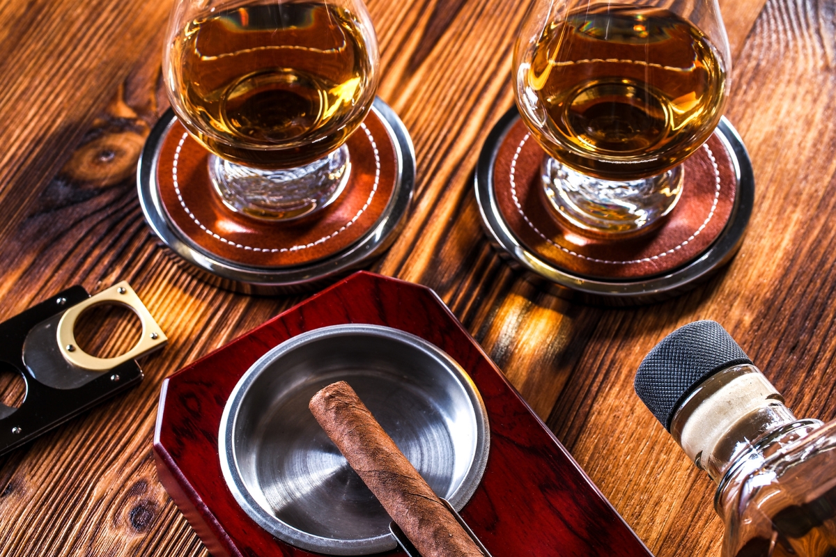 Two glasses of amber-colored liquor, a cigar in an ashtray, a cigar cutter, and a whiskey bottle on a wooden table, perfect for an evening of cigar networking.