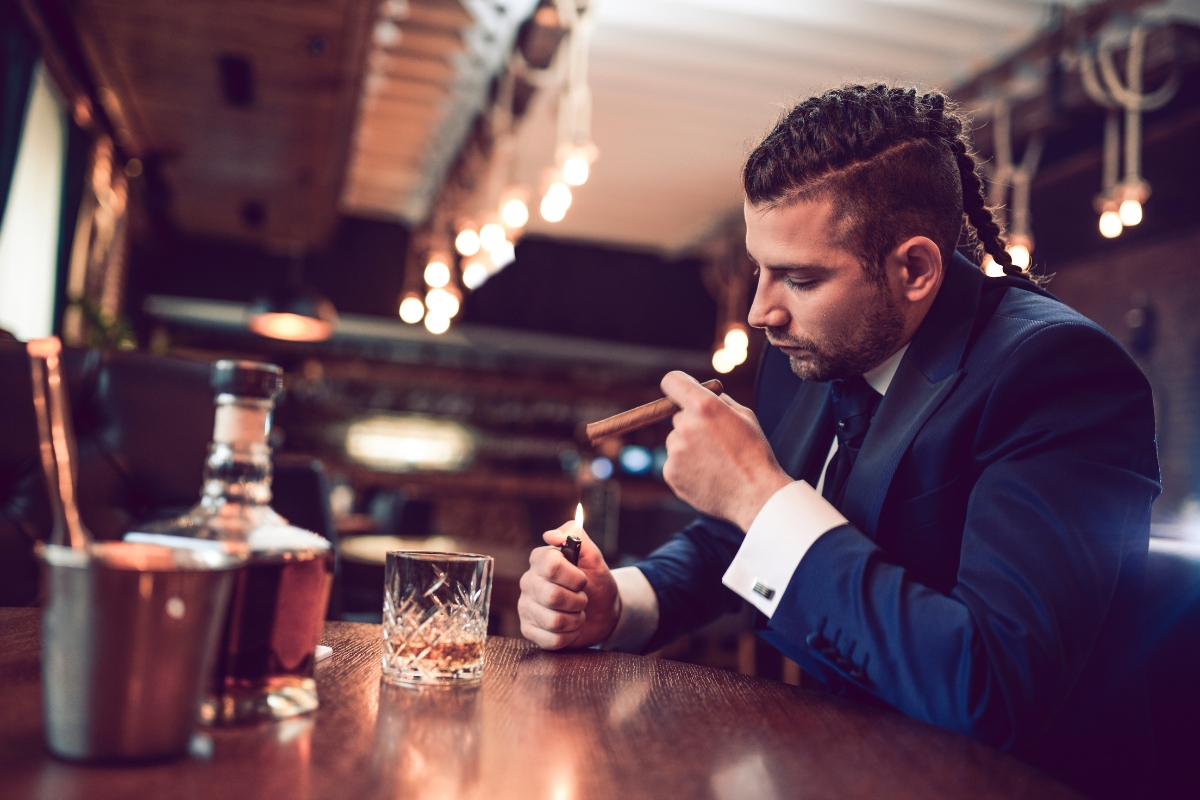 A man in a suit lights a cigar while sitting at a bar, effortlessly blending into the ambiance of high-end networking. A glass, bottle, and ice bucket are on the counter.