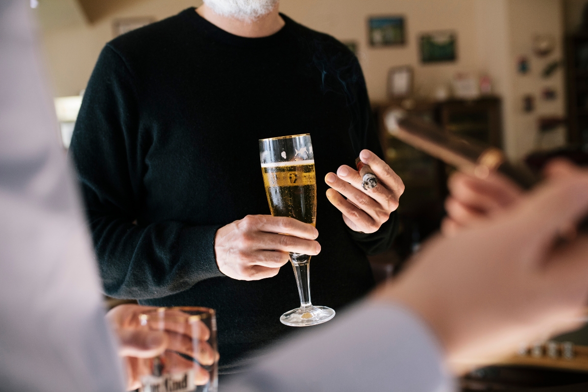 A person in a black sweater holds a glass of beer and a cigar, engaged in casual networking, with two other people holding drinks and cigars in the foreground.