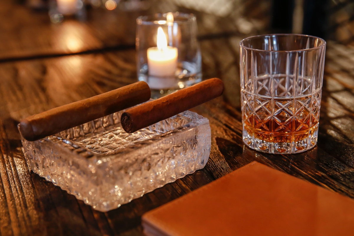 Glass of whiskey on a wooden table next to two cigars resting on an ashtray, with a lit candle in the background, setting the perfect mood for an evening of cigar networking.