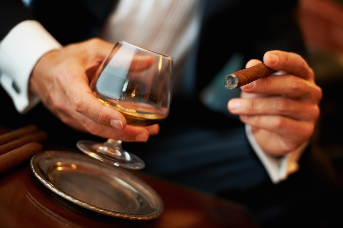 A person in a suit holds a glass of amber-colored liquid and a lit cigar, perfect for networking, with a silver tray on the table nearby.