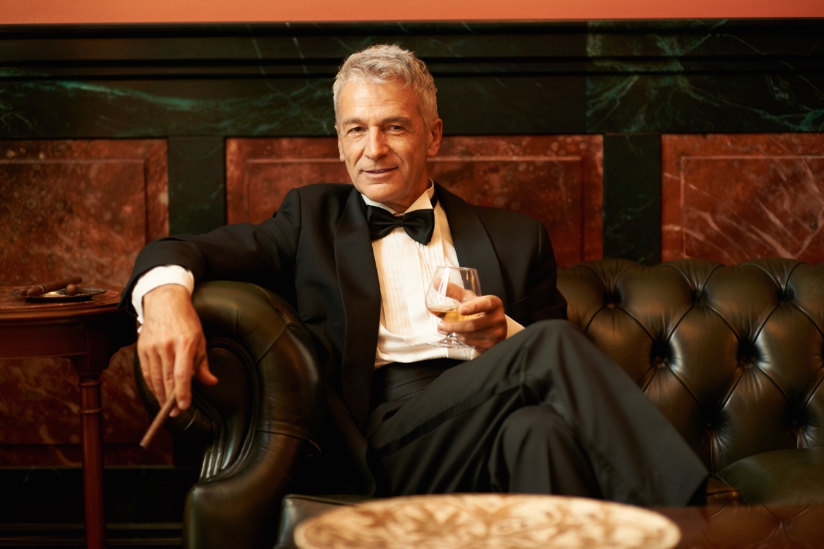 A man in a black tuxedo sits on a leather sofa holding a glass of whiskey in one hand and a cigar in the other, contemplating his next networking move.