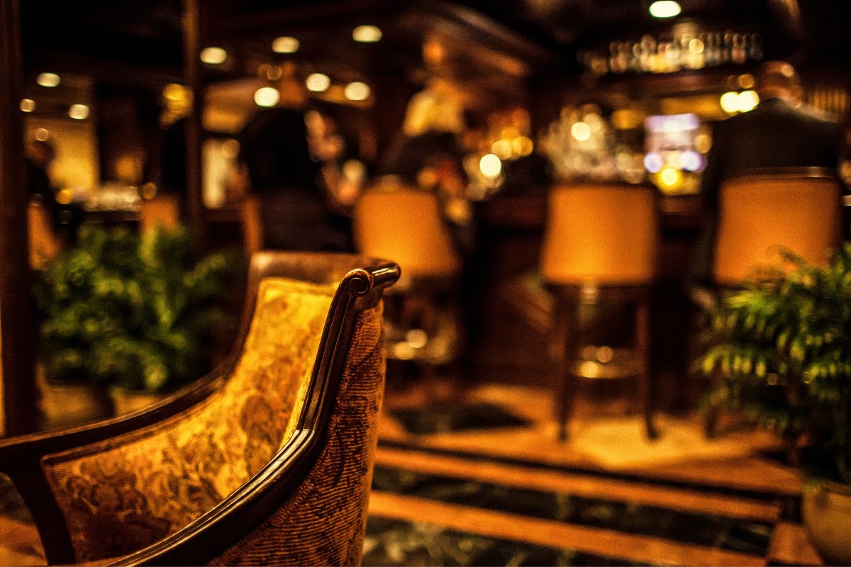 A dimly lit bar scene with a focus on an upholstered chair in the foreground and patrons practicing cigar lounge etiquette while sitting at the bar in the background.