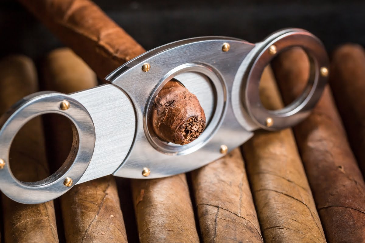 A close-up image of a steel cigar cutter resting on several brown cigars, perfect for mastering Cigar Lounge Etiquette.