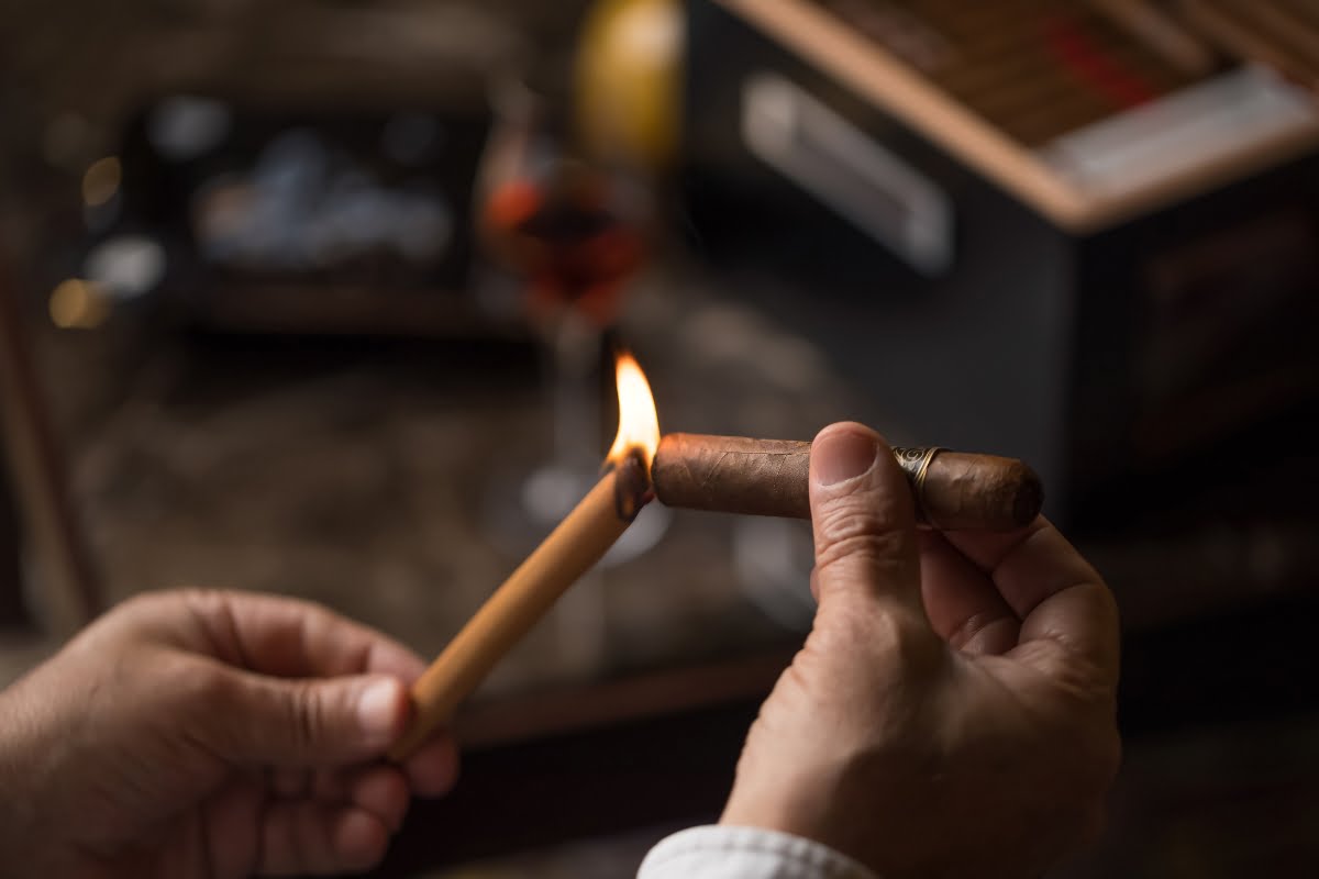 A person is holding a cigar while lighting it with a wooden stick, showcasing impeccable Cigar Lounge Etiquette. A glass of drink and a storage box are in the blurred background.