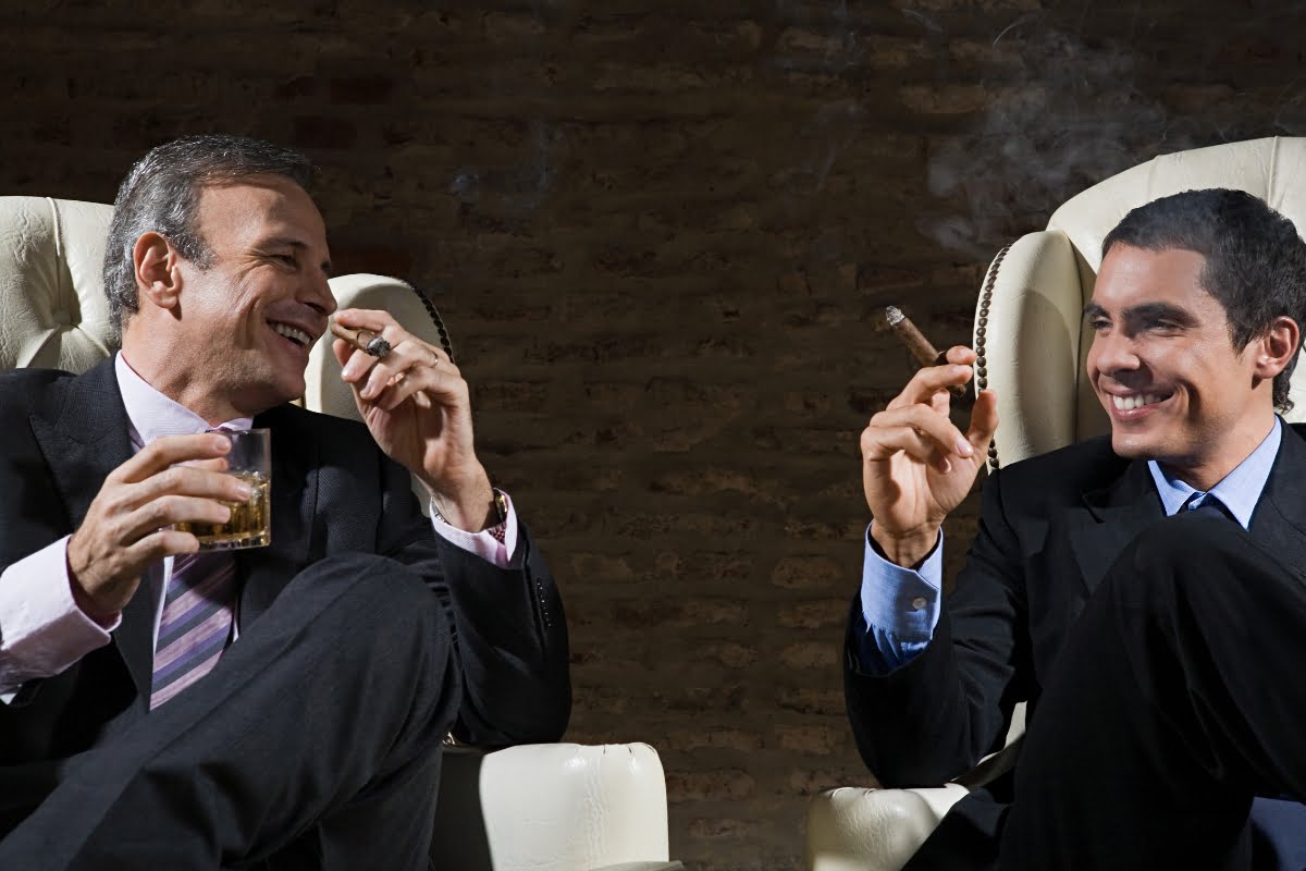 Two men in suits are sitting in armchairs, smiling and holding cigars, embodying the essence of cigar lounge etiquette. One man is holding a glass of amber liquid, possibly whiskey. The background is a brick wall.