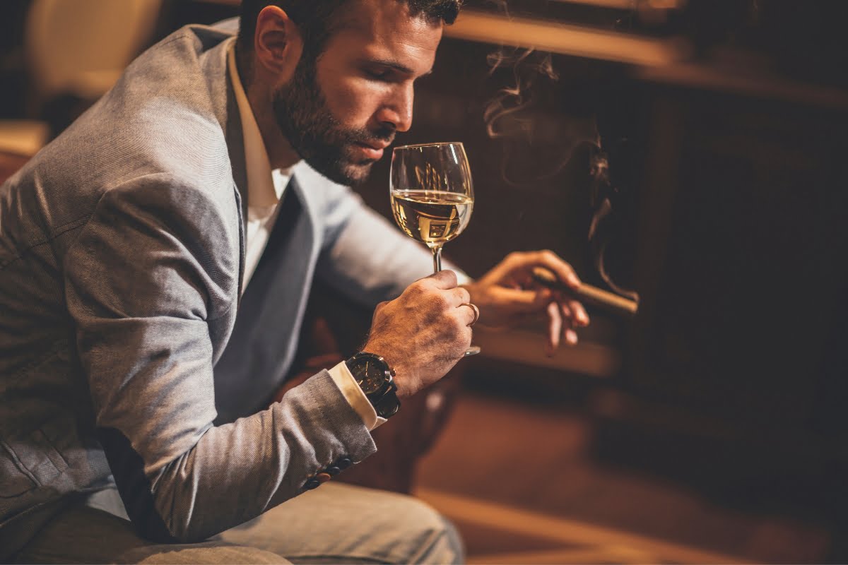 A man in a grey blazer sits indoors, adhering to Cigar Lounge Etiquette, holding a glass of white wine in one hand and a smoking cigar in the other.