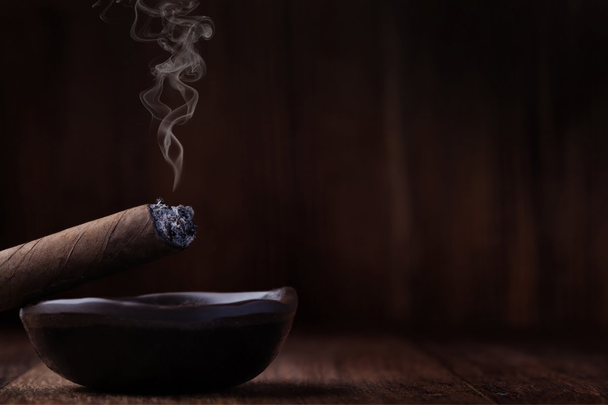 A lit cigar rests on a dark ashtray, with smoke curling upwards against a dark wooden background, embodying the essence of refined cigar lounge etiquette.