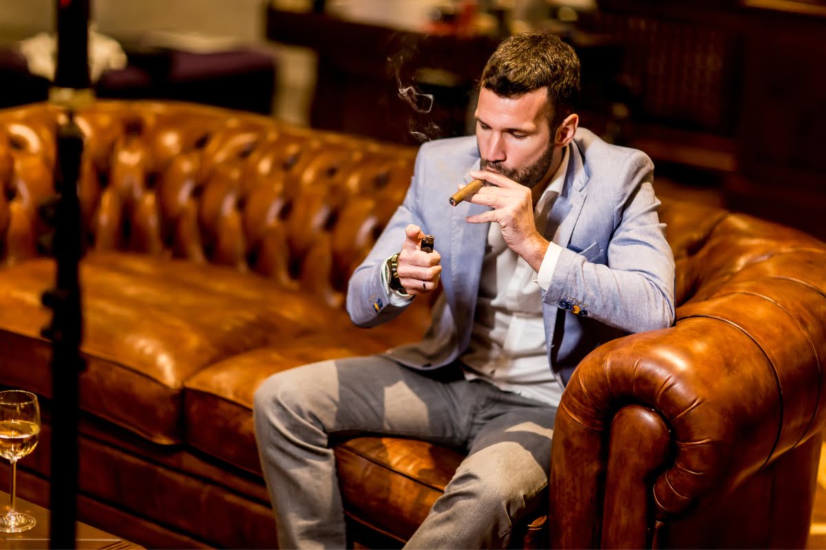 In adherence to Cigar Lounge Etiquette, a man in a light blue blazer sits on a leather couch, lighting a cigar with a lighter.