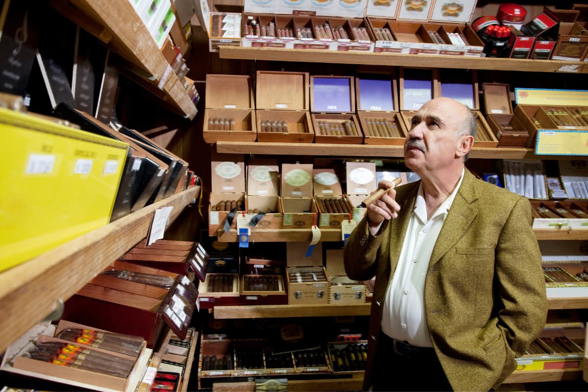 A man in a brown blazer stands in a cigar shop, holding a cigar and observing the impeccable organization of various cigars on wooden shelves, embodying the essence of refined Cigar Lounge Etiquette.