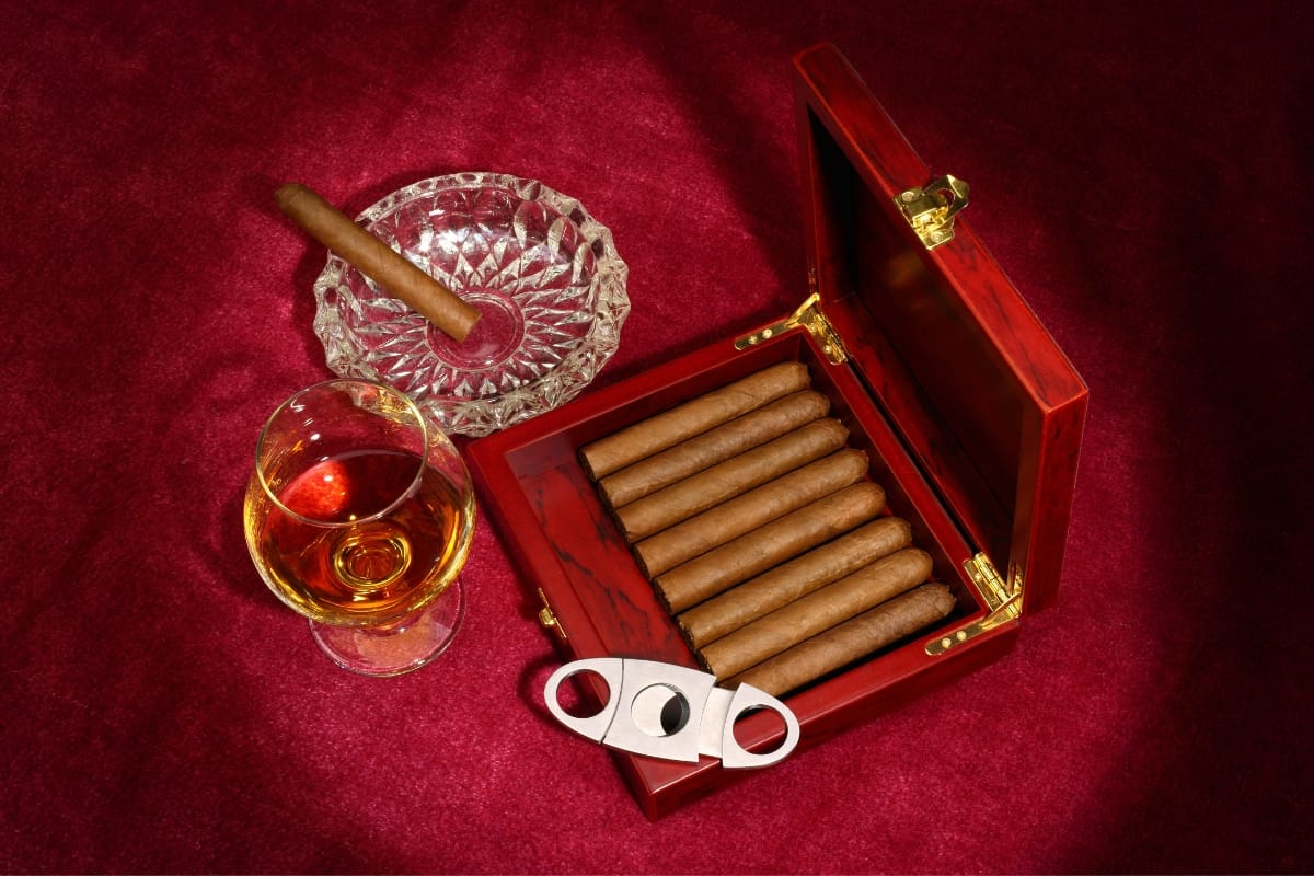 A wooden cigar box containing several gourmet cigars, an ashtray with a lit cigar, a glass of amber liquid, and a cigar cutter are elegantly arranged on a red velvet surface.