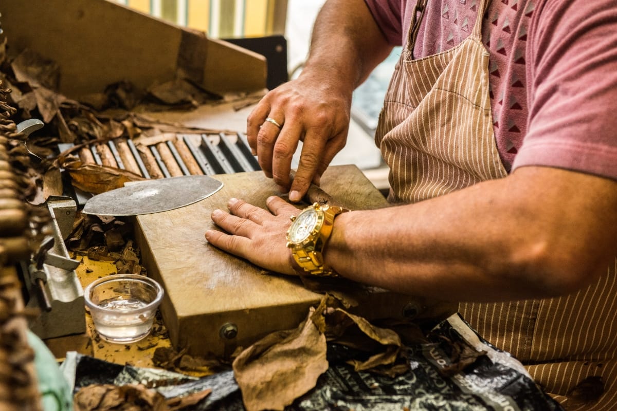 A person wearing an apron and a gold watch carefully rolls a gourmet cigar on a wooden board, surrounded by tobacco leaves and tools.