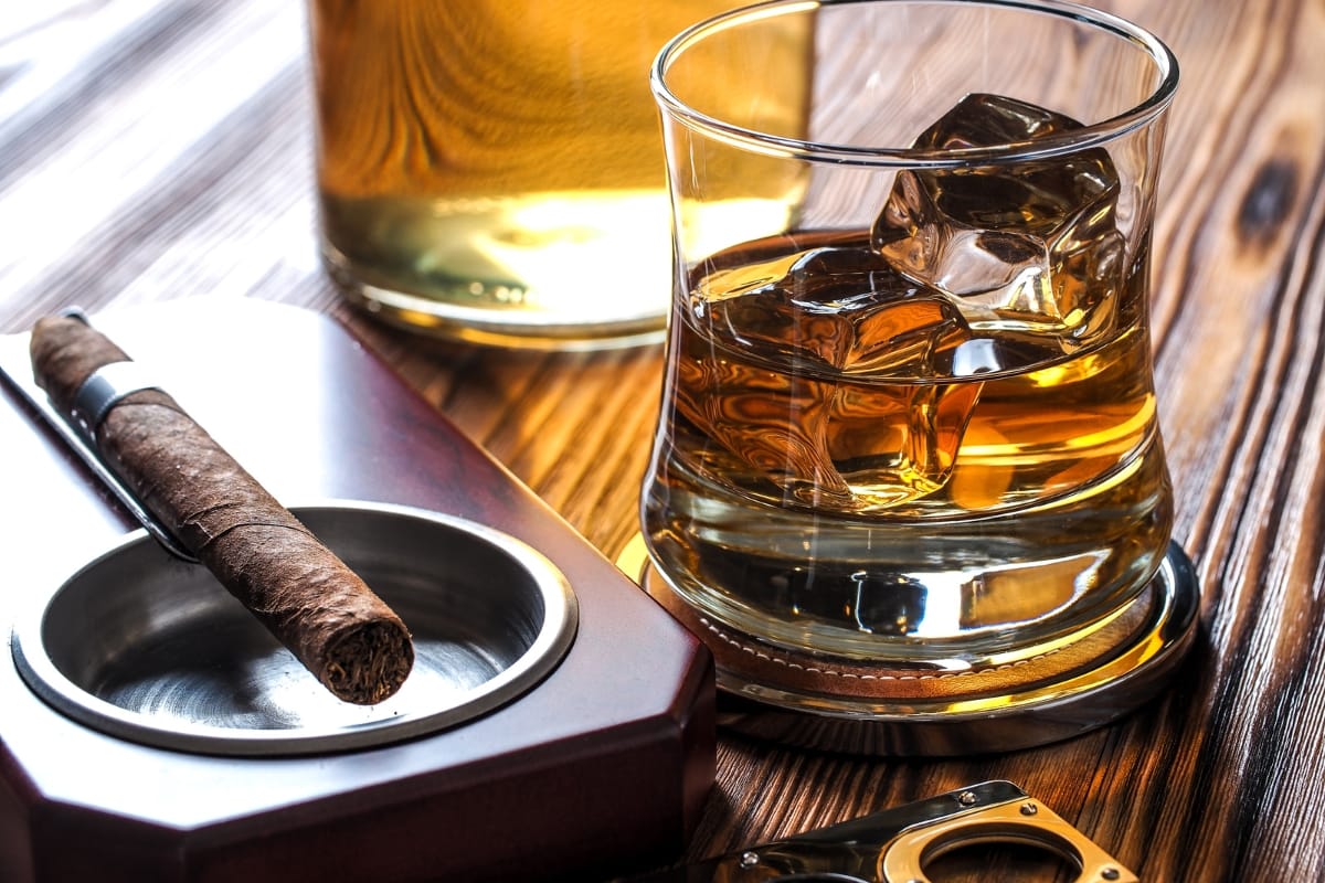 A glass of whiskey with ice, a gourmet cigar in an ashtray, and a bottle of whiskey on a wooden table.