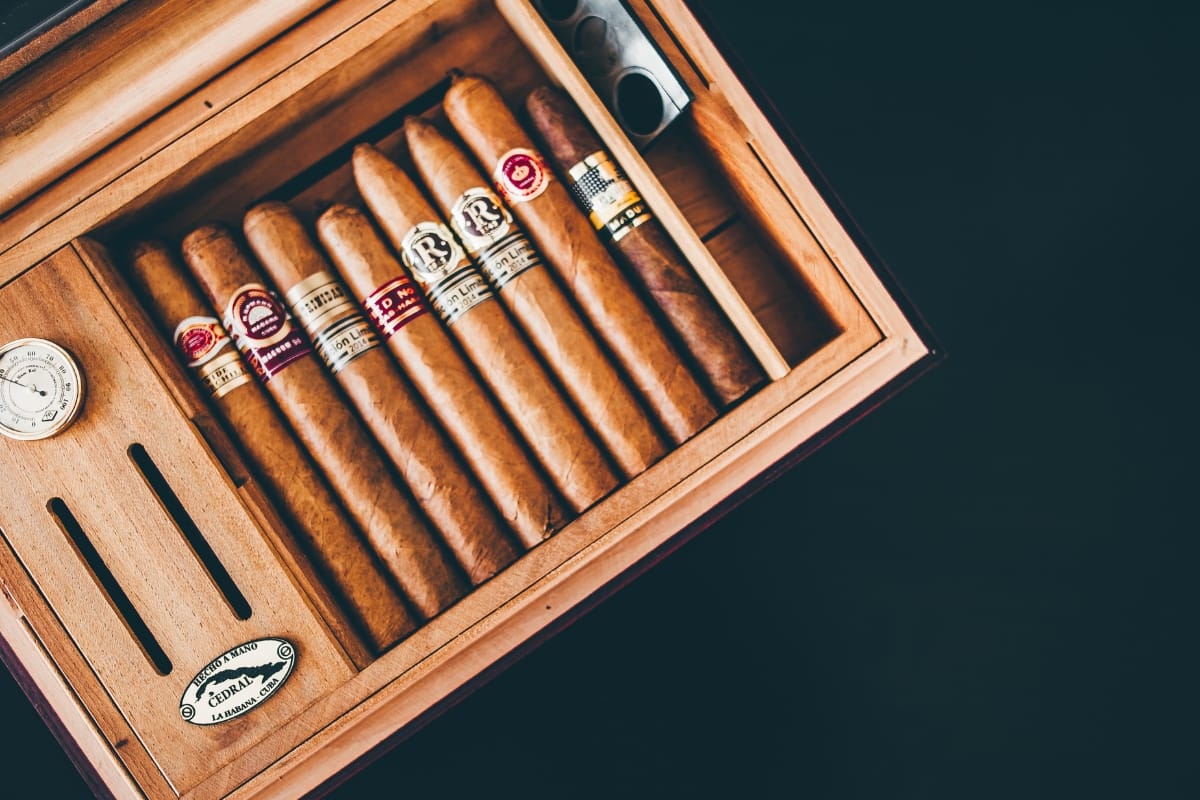 A wooden humidor containing seven gourmet cigars, a hygrometer on the left, and a metal cigar cutter in the top right corner.