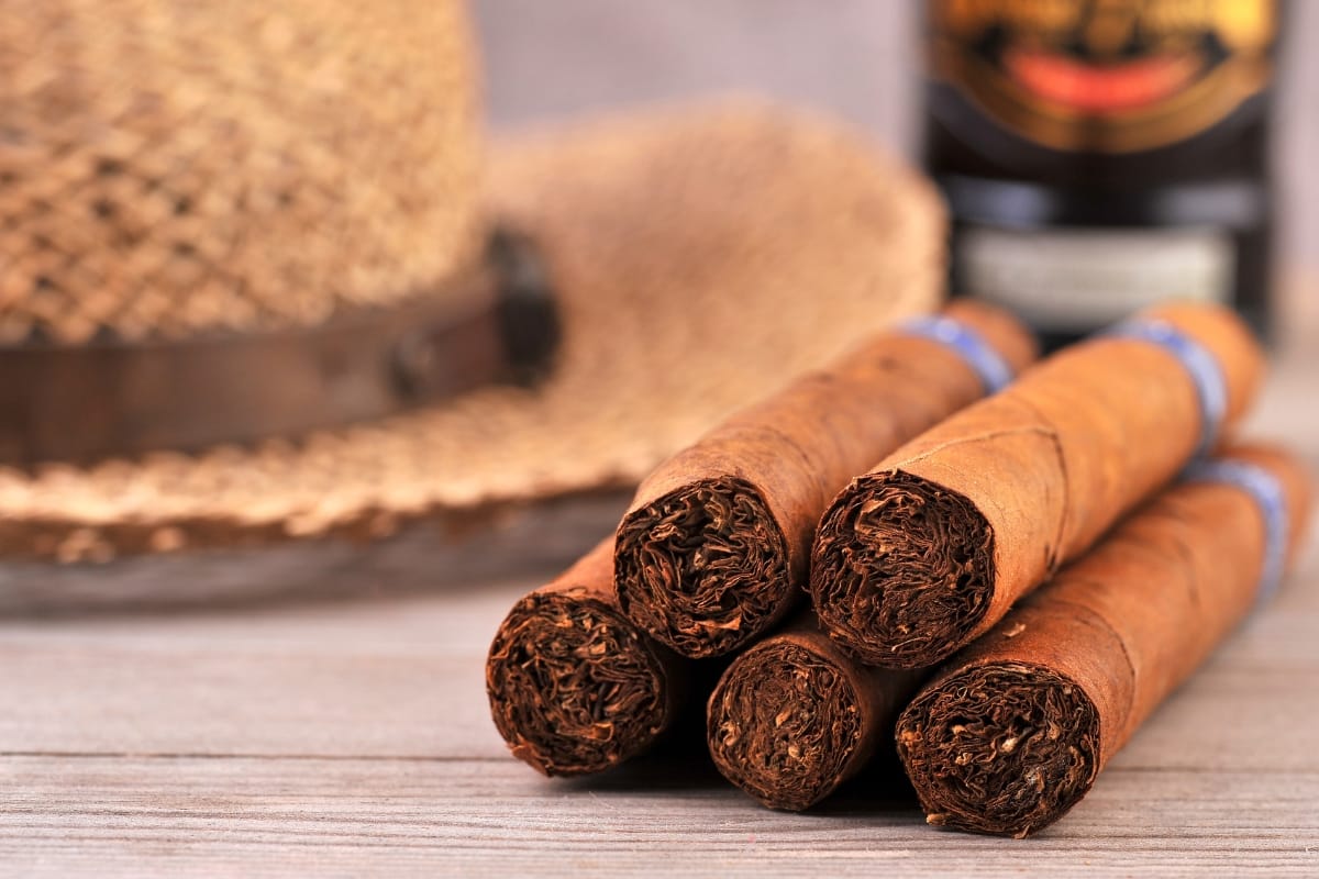 A close-up of six gourmet cigars placed on a wooden surface with a blurred straw hat and bottle in the background.