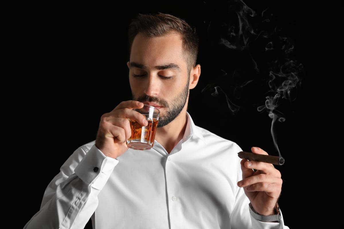 A man in a white shirt holds a glass of fine spirits and a lit cigar against a dark background.