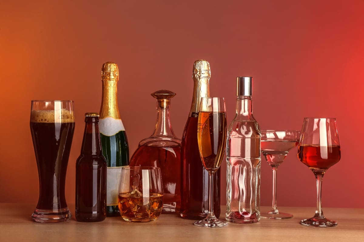 A variety of alcoholic beverages in bottles and glasses, including beer, wine, and spirits, arranged on a table against a red background, with a fine cigar adding an air of sophistication.