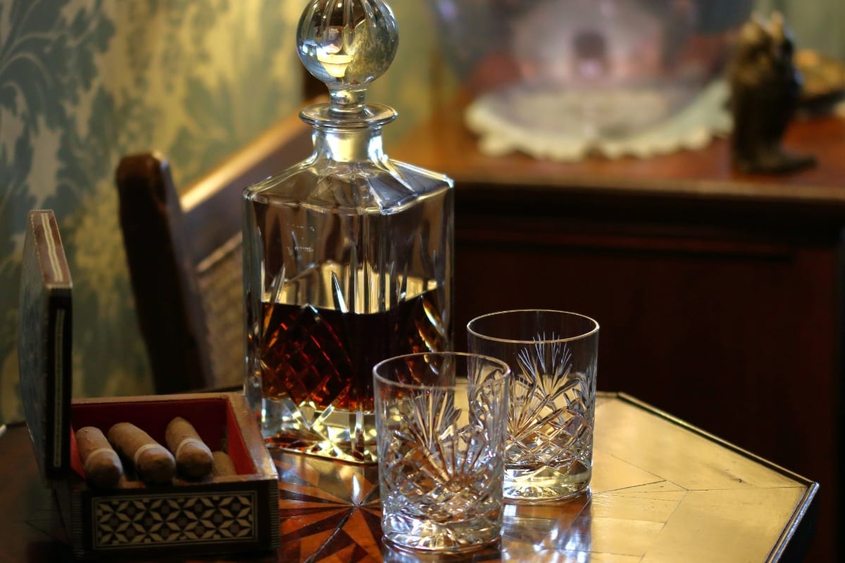 A crystal decanter brimming with fine spirits and two empty glasses sit elegantly on the wooden table, accompanied by an open box with cigars, enticing a moment of indulgence.