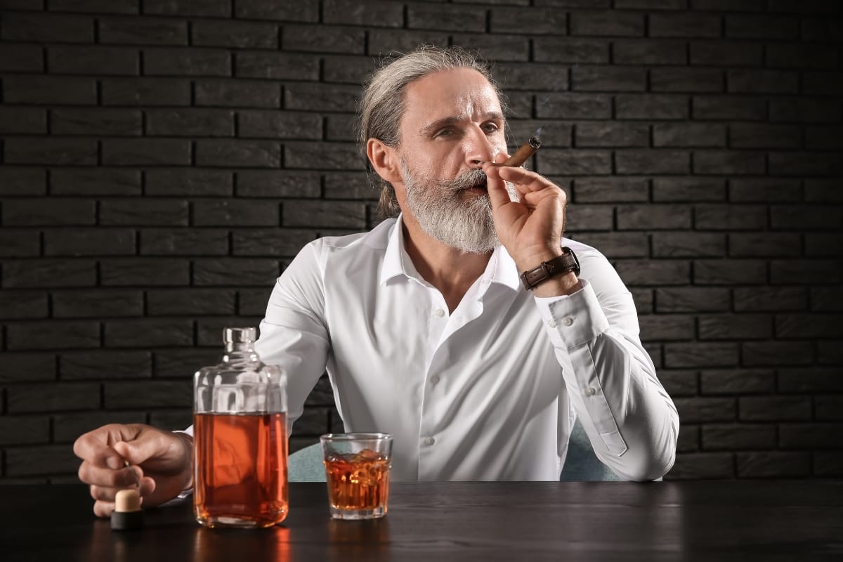 A man with gray hair and beard sits at a table, wearing a white shirt, savoring a cigar. A bottle of fine spirits and a glass filled with the dark liquid are on the table. A brick wall forms the backdrop.