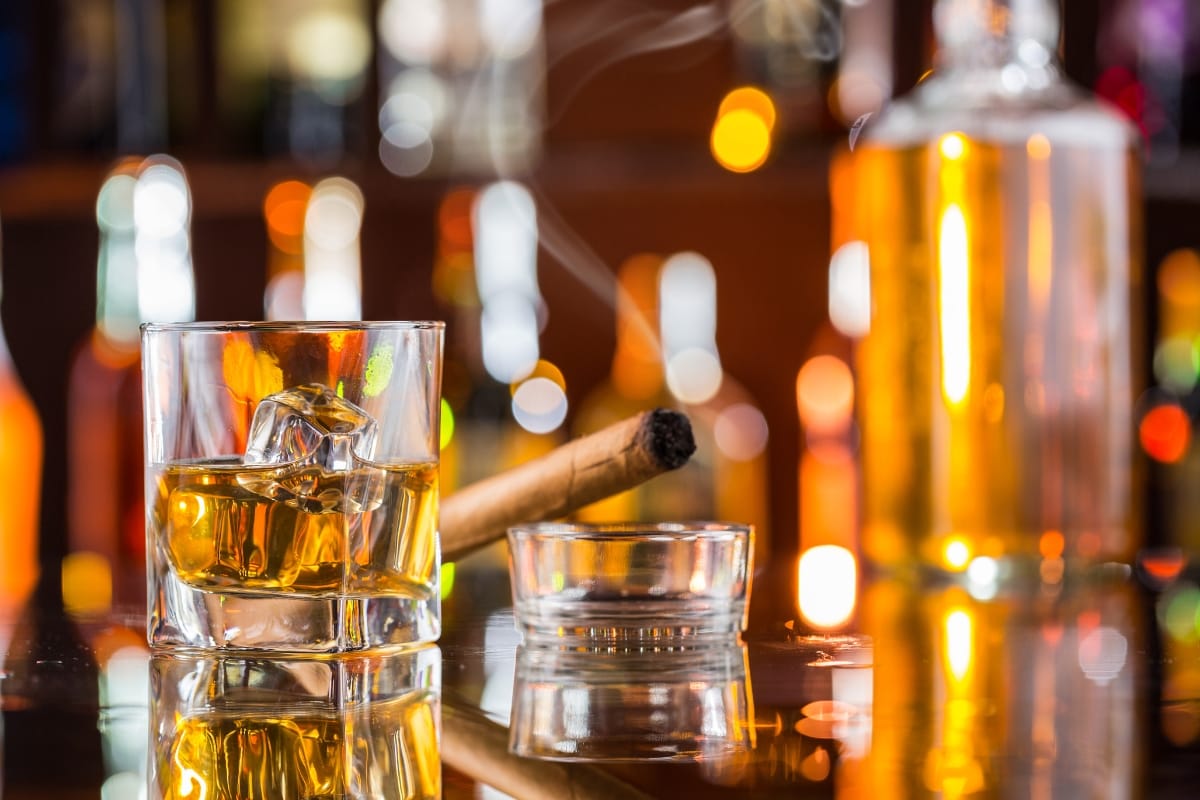 A glass of whiskey with ice sits next to a lit cigar in an ashtray, accompanied by blurred bottles of spirits in the background.