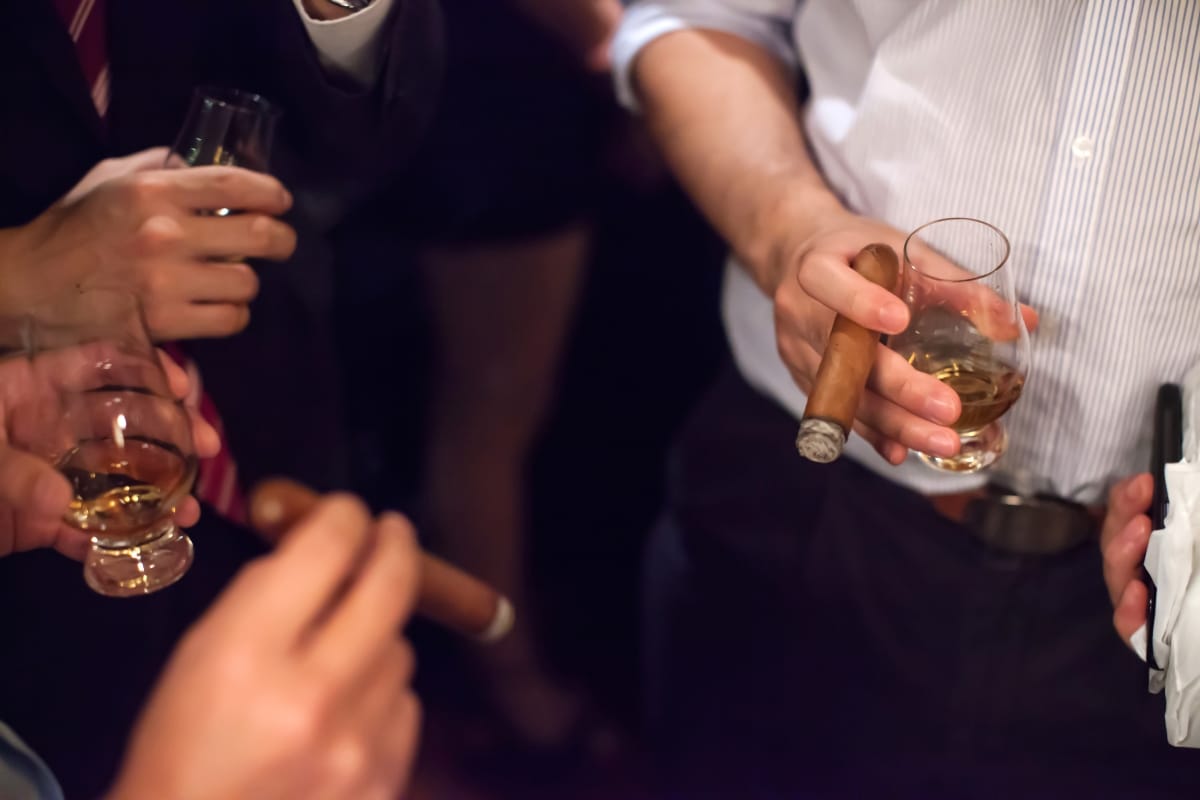 Amidst a dimly lit setting, people savor cigars and spirits, their glasses of whiskey catching the soft glow of the ambient light.