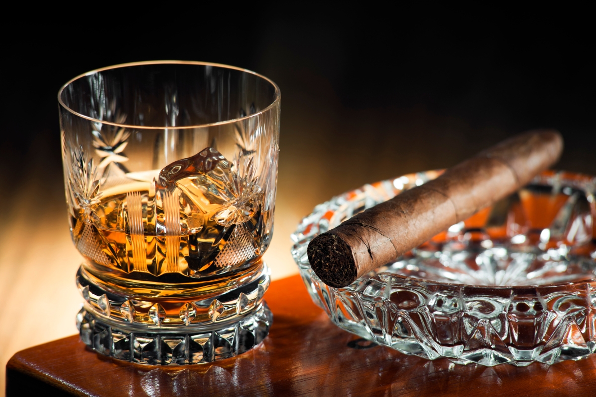 A glass of fine spirits with ice sits next to a cigar resting on a glass ashtray on a wooden surface.