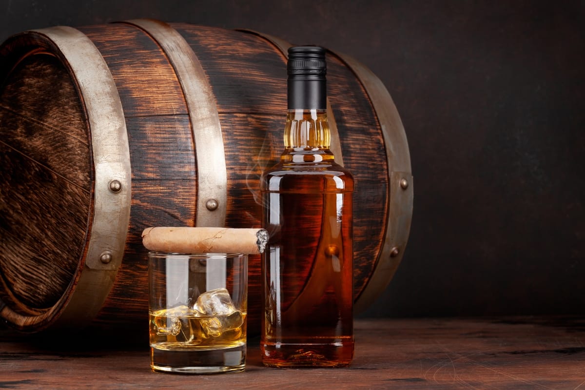 A bottle of rich spirits sits next to a glass with ice, while a cigar rests on the wooden surface, evoking an atmosphere of classic indulgence. In the background, a wooden barrel completes this picture of refined elegance.