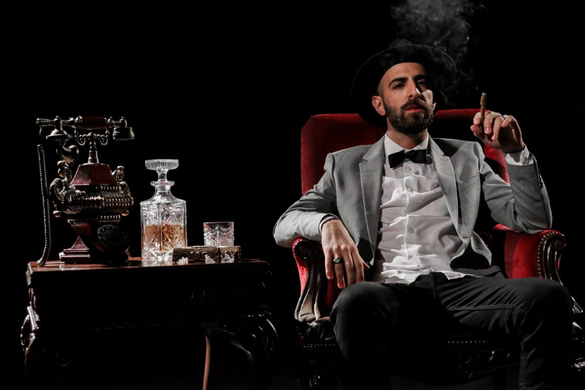 A man in a suit and hat epitomizes the cigar lifestyle as he relaxes in a red armchair, savoring a cigar. A vintage telephone and whiskey decanter on the side table complete the sophisticated scene.