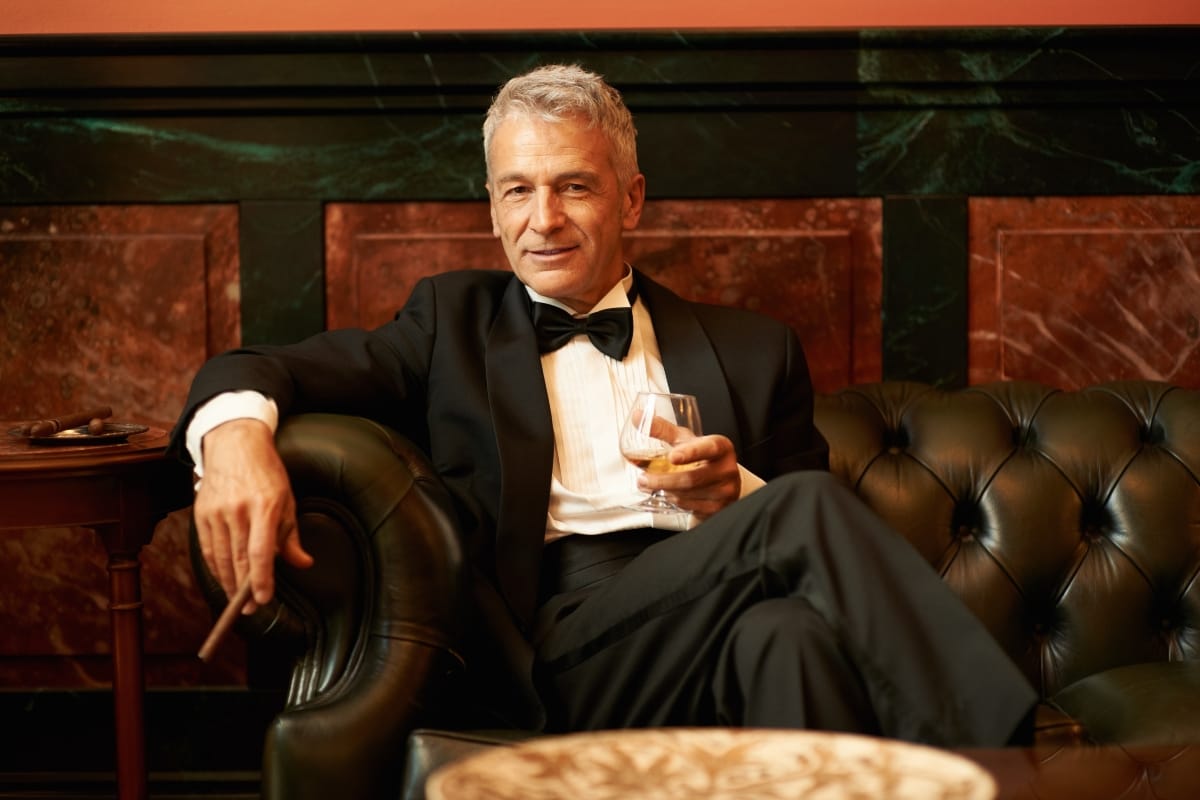 A man encapsulating the cigar lifestyle sits on a luxurious leather couch, dressed in a tuxedo, savoring both his drink and a fine cigar.