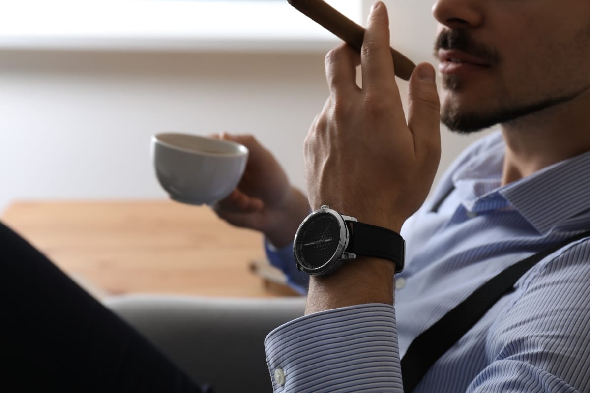 A person in a striped shirt and suspenders embodies the cigar lifestyle as they hold both a coffee cup and a cigar. Adorned with a wristwatch, they seem relaxed and content seated indoors.