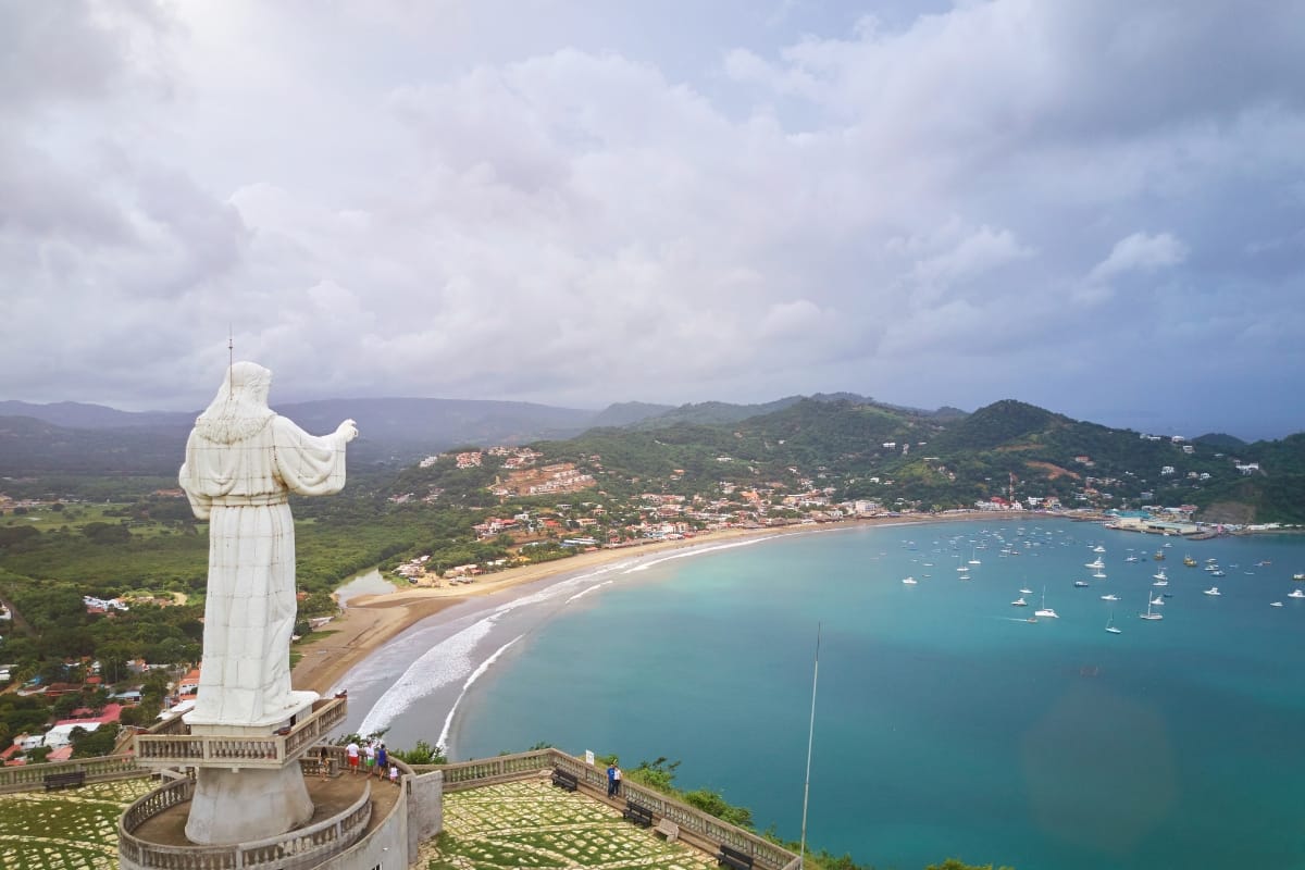 Overlooking a coastal town with a curved beach, lush hills, and numerous boats in the bay under a cloudy sky, this statue marks one of the travel destinations for cigar lovers seeking serene views and rich experiences.