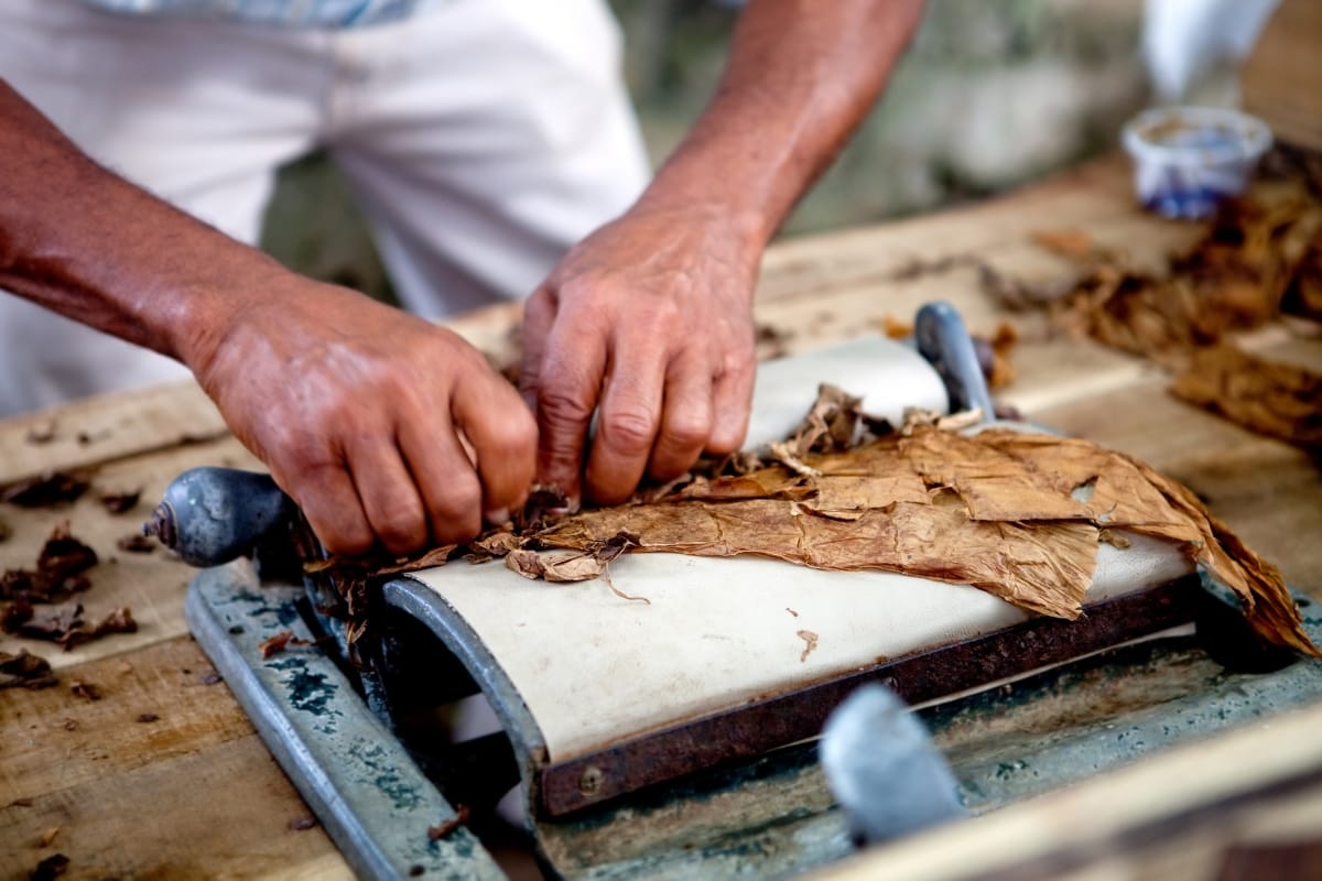 Hands rolling a cigar on a wooden table using a metal tool, crafting flavors perfect for travel destinations for cigar lovers.