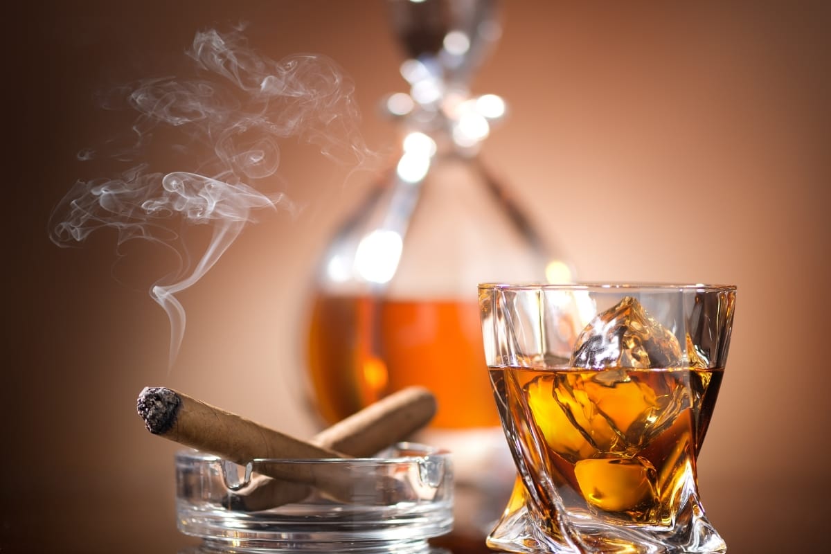 A glass of whiskey with ice sits next to a lit cigar in an ashtray, evoking the charm of travel destinations for cigar lovers. A decanter is blurred in the background.