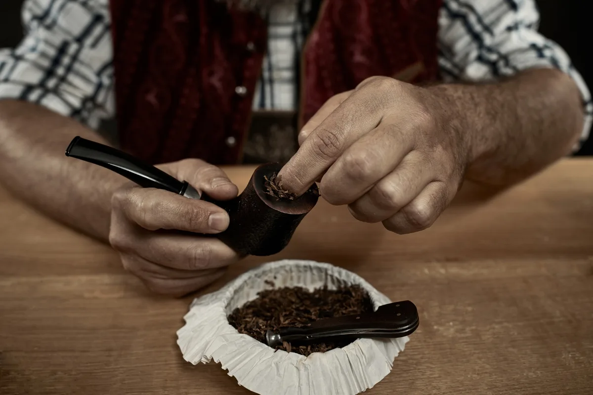 On a wooden surface, someone carefully fills a tobacco pipe with loose tobacco, while a tamper tool lies nearby, ready for use in this ritual of tobacco pipe care.