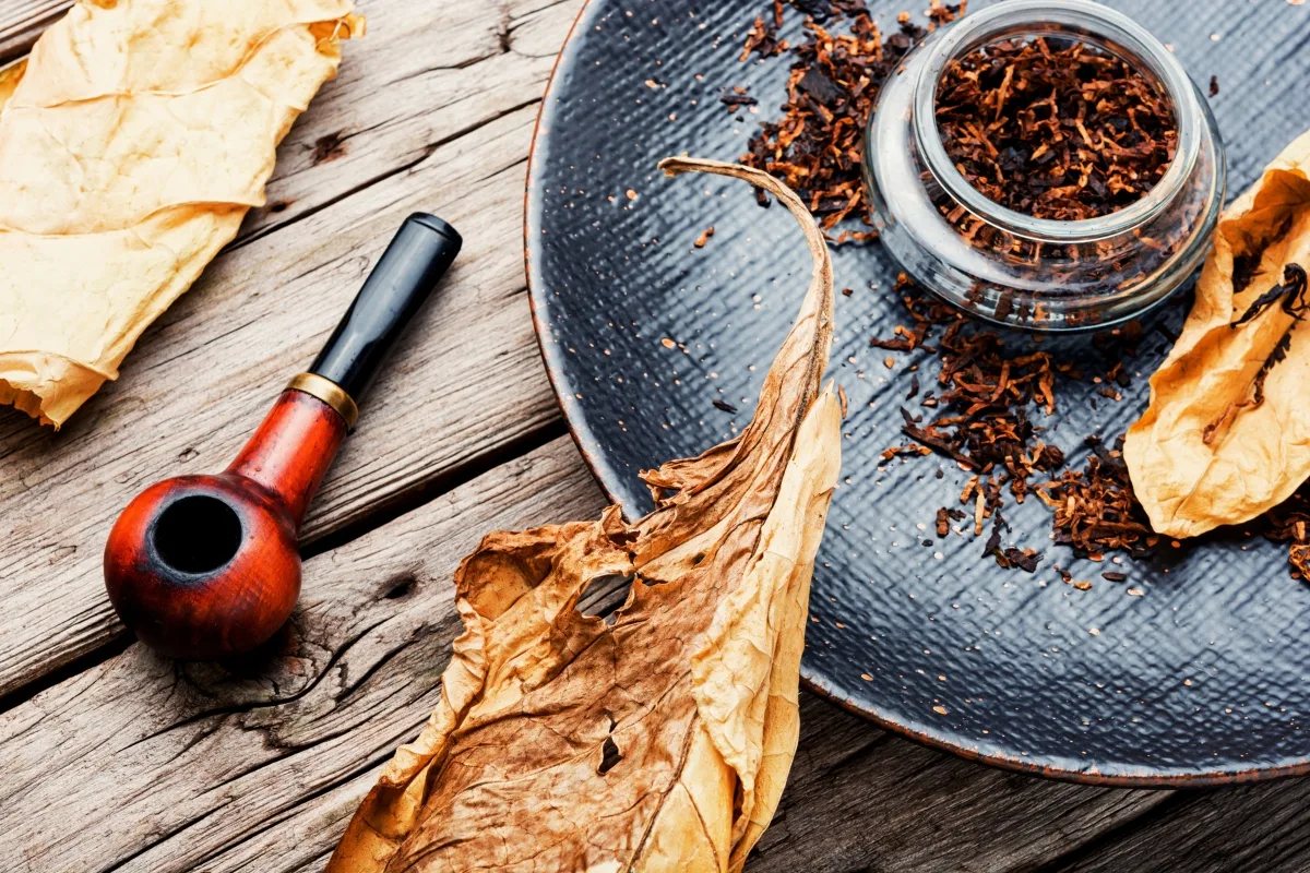 Wooden smoking pipe and dried tobacco leaves rest on a rustic wooden surface, with a glass jar containing tobacco placed on a round ceramic plate, exemplifying the finest in tobacco pipe care.