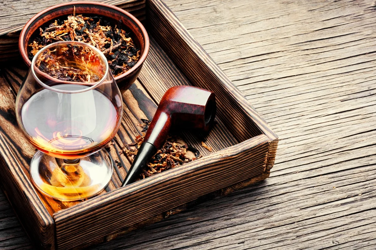 A wooden tray holds a glass of amber liquid, a well-maintained smoking pipe, and a bowl of loose tobacco on a textured wooden surface.