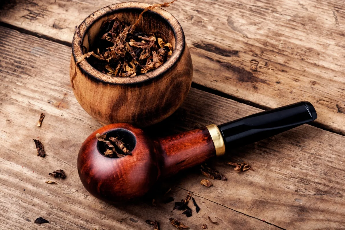 A wooden tobacco pipe and a bowl filled with dried tobacco sit on a rustic wooden surface, highlighting the timeless debate of wooden vs clay tobacco pipes.