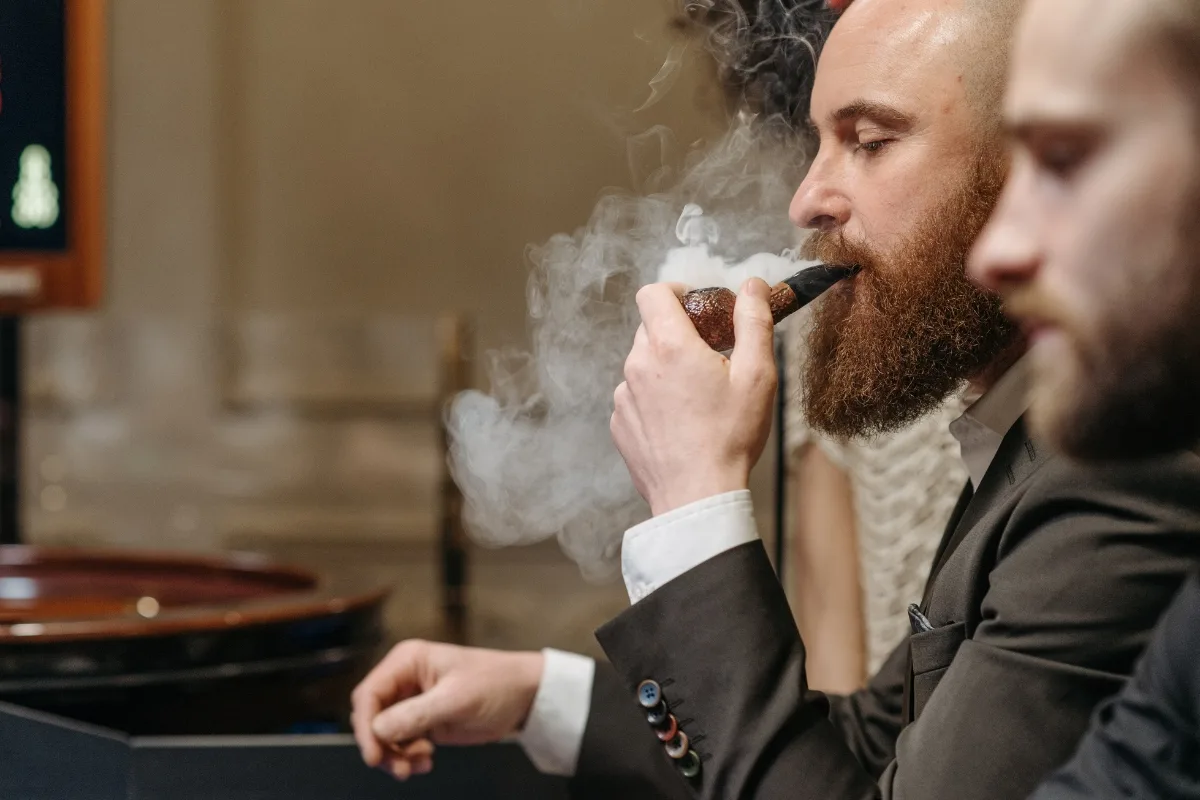 A man in a suit smokes a pipe indoors, with another gentleman by his side. The swirling smoke highlights the elegance of his wooden tobacco pipe, contrasting with the subtle charm of his companion's clay version.