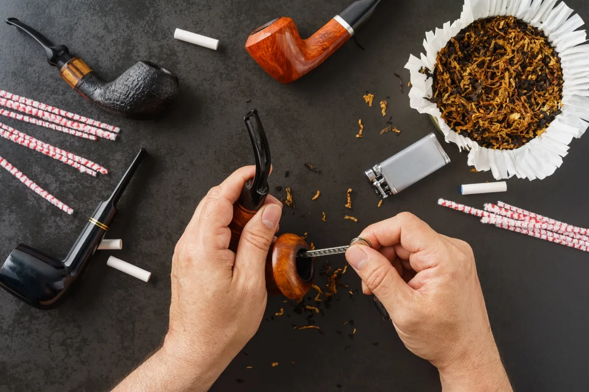 Hands filling a tobacco pipe with shredded tobacco on a dark surface; multiple pipes, including wooden and clay varieties, a lighter, and rolled filters are visible.