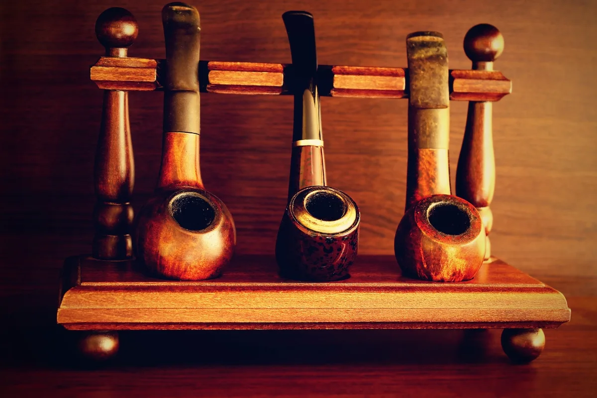A wooden pipe rack holds four tobacco pipes, each uniquely designed, against a rustic wooden background. Whether you prefer the classic charm of wooden or the traditional appeal of clay tobacco pipes, this elegant display captures the artistry of fine smoking instruments.