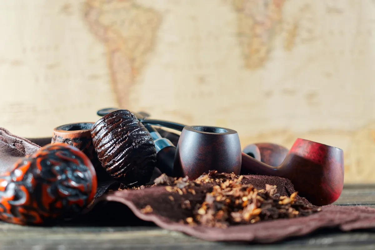 Assorted wooden and clay tobacco pipes along with loose tobacco rest on a brown cloth, set against the backdrop of a blurred world map.